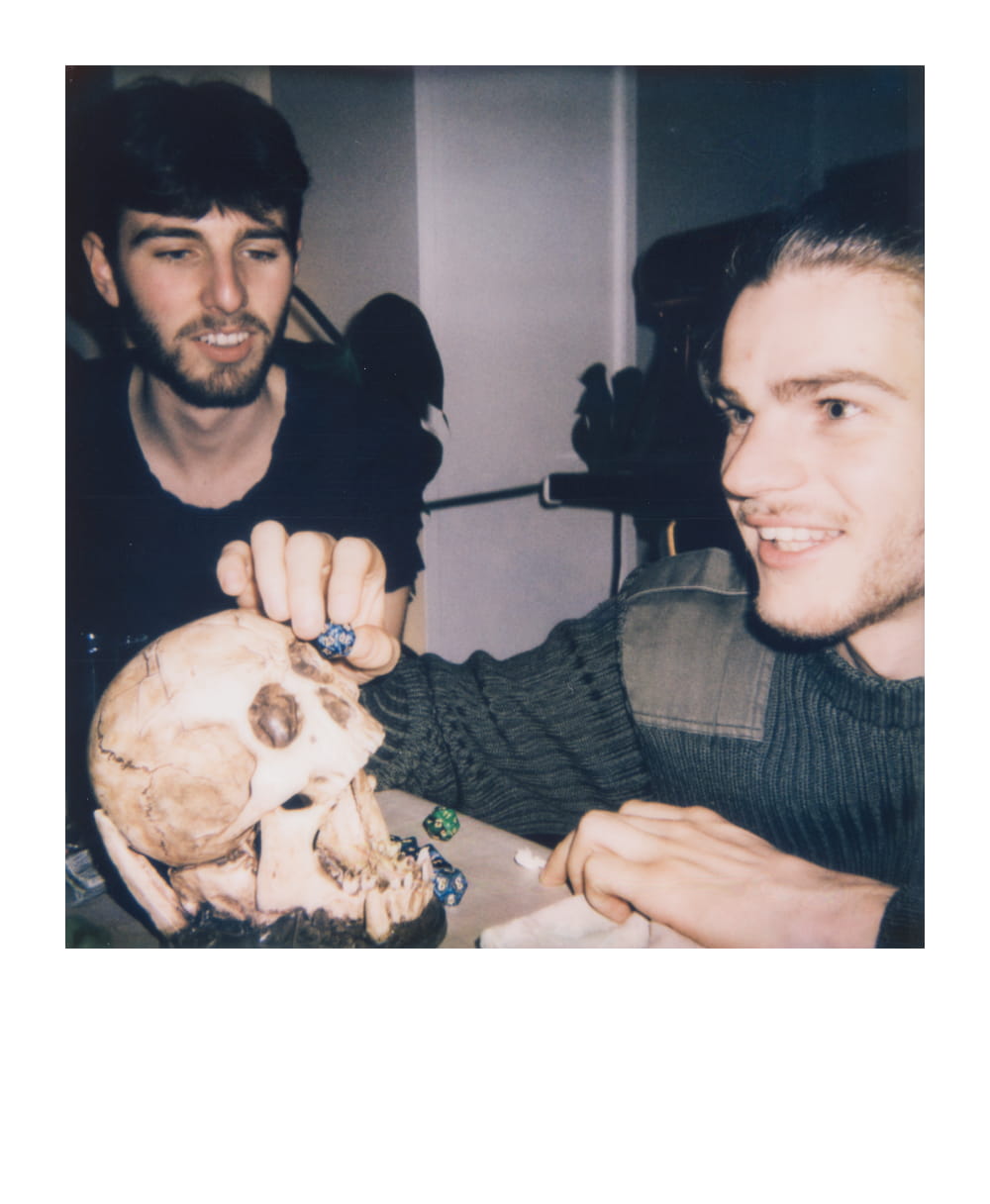 Two friends playing D&D at a table with dice and a human skull prop, deep in conversation during their role-playing game.