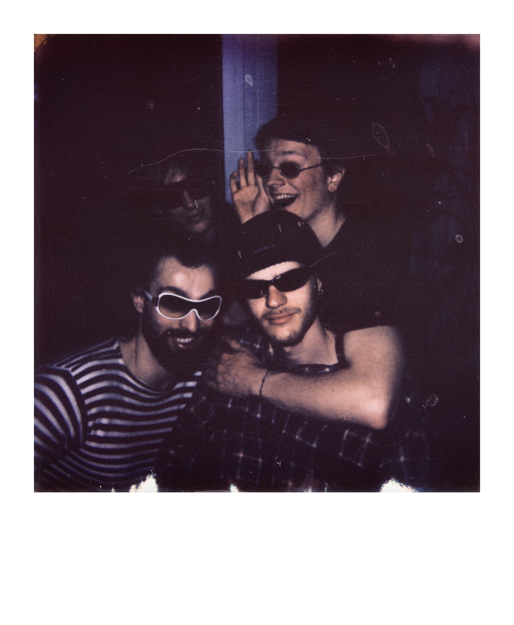 Group of four friends in playful sunglasses and dark outfits, posing indoors with a fun and casual vibe.