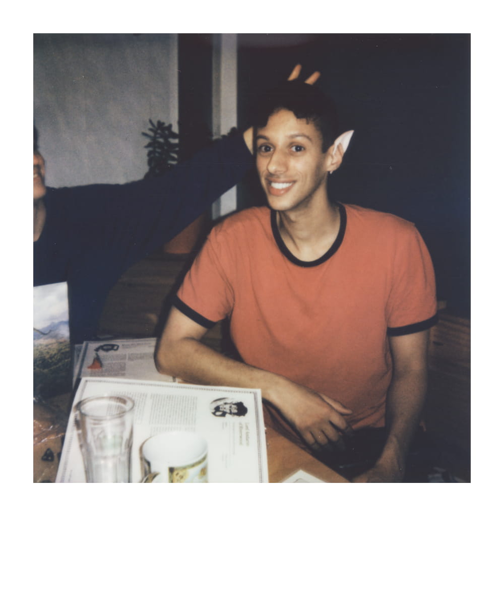 A player wearing elf ears smiling at the camera, sitting at a table with D&D character sheets and dice, immersed in the game.